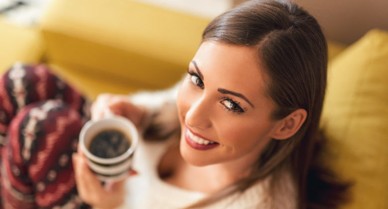 Beautiful young smiling woman relaxing on sofa at living room while enjoying in coffee time.